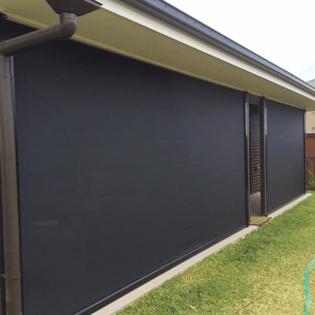 Outdoor black roller blinds fully cover the windows of a house with beige walls, adjacent to a green lawn and a garden hose.