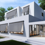 A modern white two-story house with large windows covered by gray exterior awnings, a patio with chairs and table, and a green lawn.