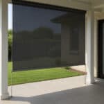 A black outdoor roller shade is lowered on a covered patio, with tiled flooring and a view of a fenced backyard and lawn.