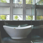 A modern bathroom with a white freestanding bathtub, two soap dispensers, a glass shower enclosure, and large windows with blinds.
