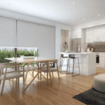 Modern open-plan kitchen and dining area with light wood floors, white cabinetry, a dining table with chairs, and roller blinds on large windows.