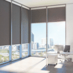 Modern office with large windows, partially covered by dark roller blinds, overlooking a cityscape with tall buildings and a sunny sky. A desk and chair are in the corner.
