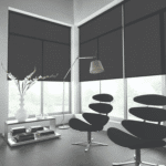 Modern living room with large dark roller shades, two contemporary black chairs, a floor lamp, a white vase with flowers, and a table stacked with books near wide windows.