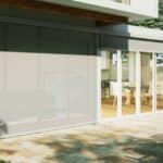 Modern patio with a dining table and chairs on stone flooring, adjacent to a house with large glass sliding doors, some covered by exterior blinds.