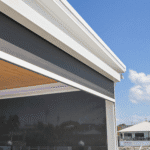 A modern building exterior with a black retractable screen or shade partially lowered, under a white roof, against a blue sky.