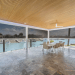 Outdoor dining area with a wooden ceiling, tiled floor, large table and chairs, overlooking a marina through clear glass barriers and roll-down shades.