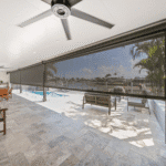 Covered patio area with ceiling fans, stone flooring, and a retractable screen overlooking a pool, outdoor seating, and waterfront view.