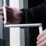 A person adjusts a white, L-shaped, folding metal arm joint with both hands near a door or window frame.