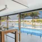 Modern outdoor patio with wooden table and pool, glass fence, palm trees, and a view of water and houses in the background.
