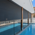 Outdoor blinds are lowered over floor-to-ceiling windows on a modern building next to a swimming pool, with blue water and glass fencing visible.
