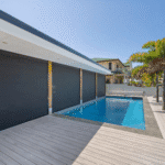 Modern outdoor swimming pool with clear water, surrounded by a light wooden deck and palm trees, adjacent to a contemporary building with large black window shades.