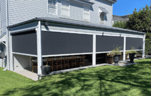 A covered patio with three large black outdoor blinds, a dining table with wooden chairs, potted plants, and a grassy yard in front.