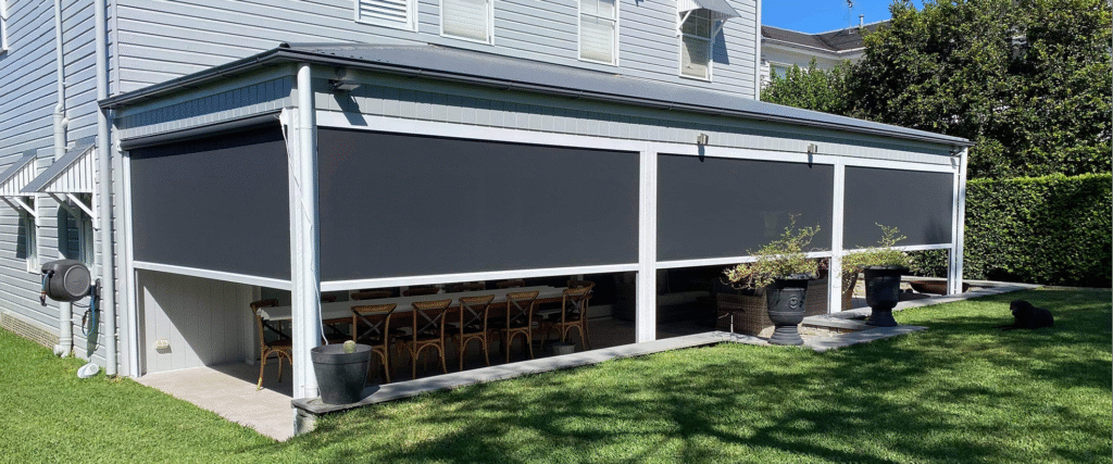 A covered outdoor patio with large black retractable screens, a long dining table with chairs, potted plants, and a grassy yard.
