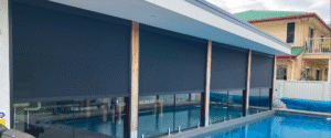 Large outdoor roller blinds cover windows along a modern poolside patio, with a glass fence and residential buildings visible in the background.
