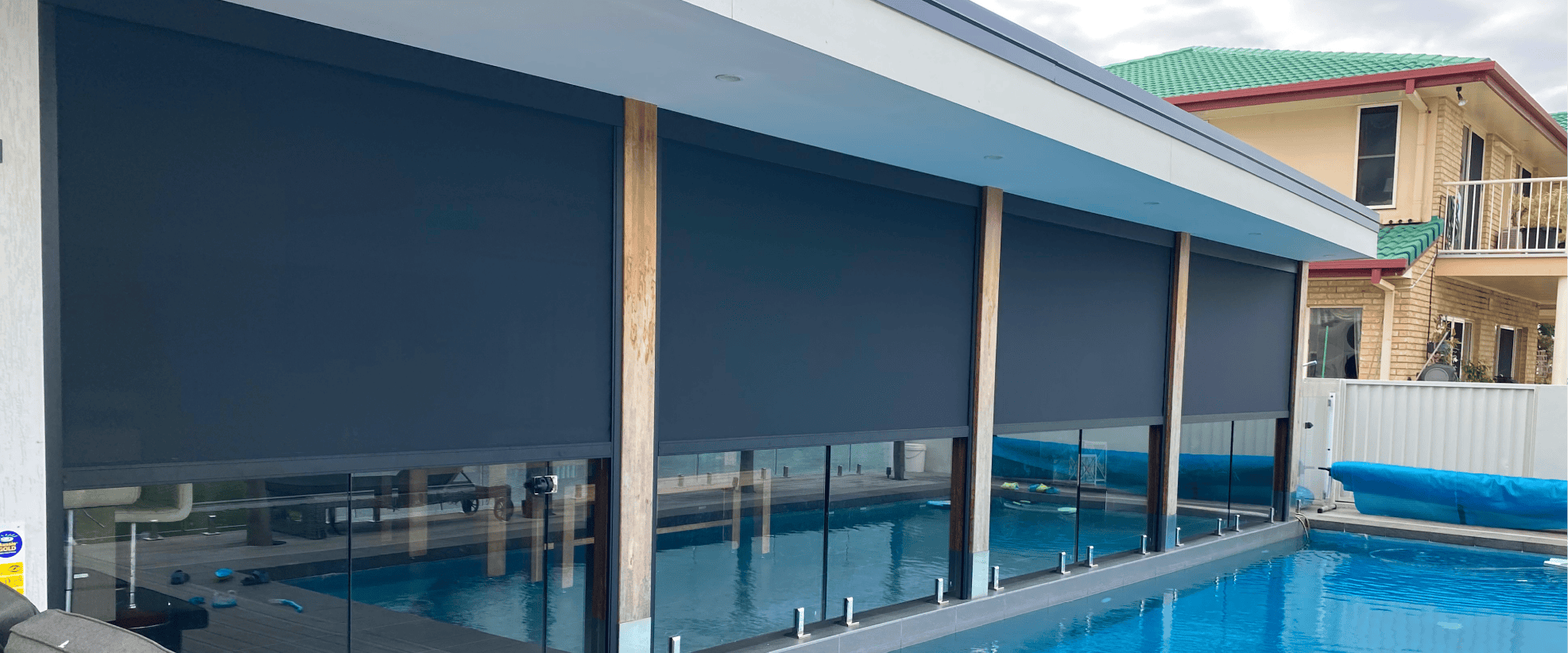 Large outdoor roller blinds cover windows along a modern poolside patio, with a glass fence and residential buildings visible in the background.