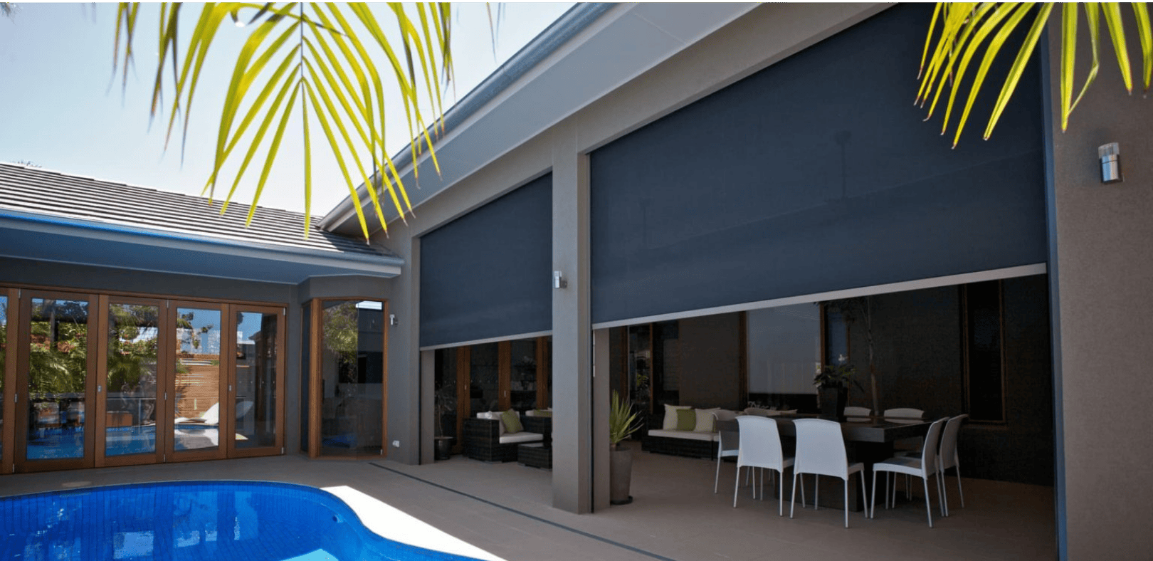 Modern patio area with a covered pool, outdoor dining set, and large retractable shade screens partially drawn down on a contemporary house. Palm fronds are visible in the foreground.