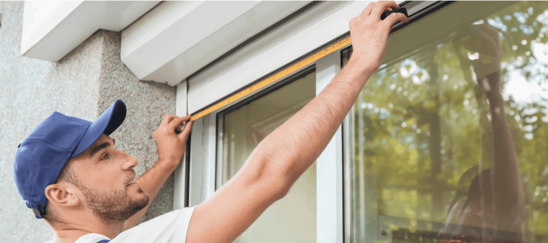 A person in a blue cap measures the width of a window using a tape measure, holding it across the top frame outside a building.