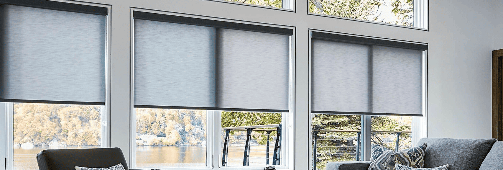 Large windows in a modern living room with light gray roller shades partially lowered, overlooking a lake and trees. A gray armchair with patterned pillows is in the foreground.
