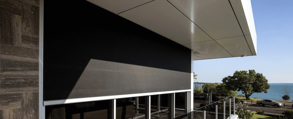 A modern building with large windows partially covered by a dark exterior screen; a balcony and water view with trees and cars in the background.