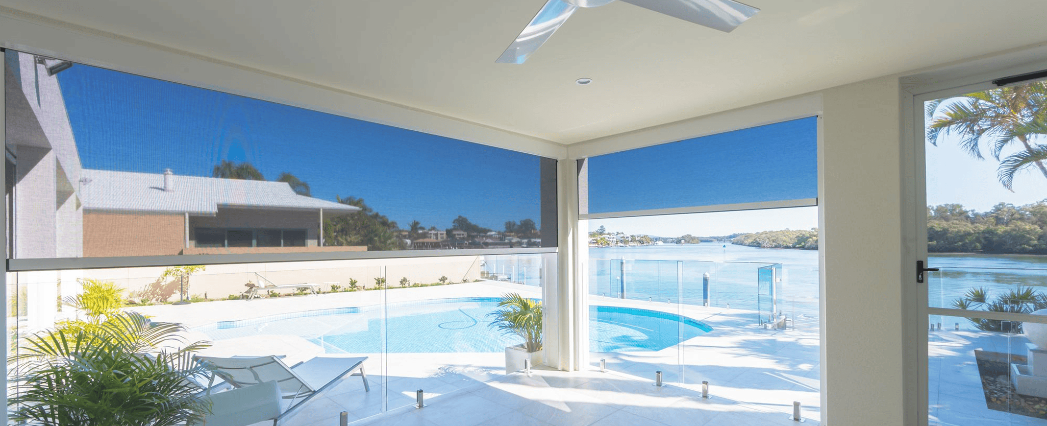 A covered patio with outdoor roller blinds overlooks a swimming pool, glass fencing, and a waterfront view on a sunny day.
