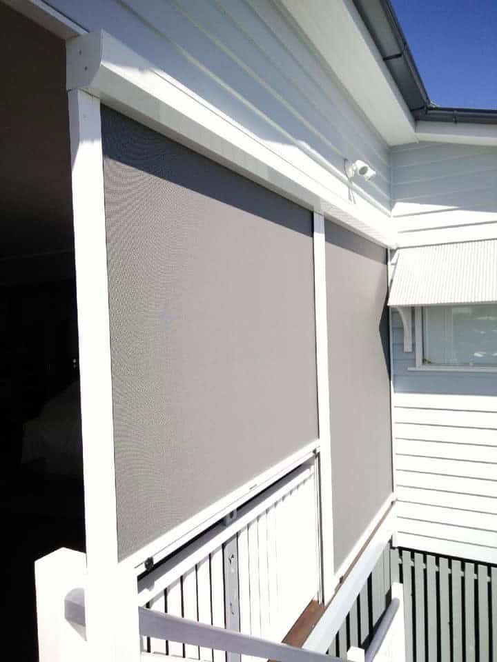 Outdoor roller blinds cover windows and veranda on a white house, providing shade and privacy. The sky is clear and blue.