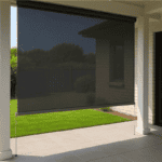A black outdoor roller shade is lowered on a covered patio, partially blocking sunlight and providing shade to the tiled area. Lawn and garden are visible beyond the shade.