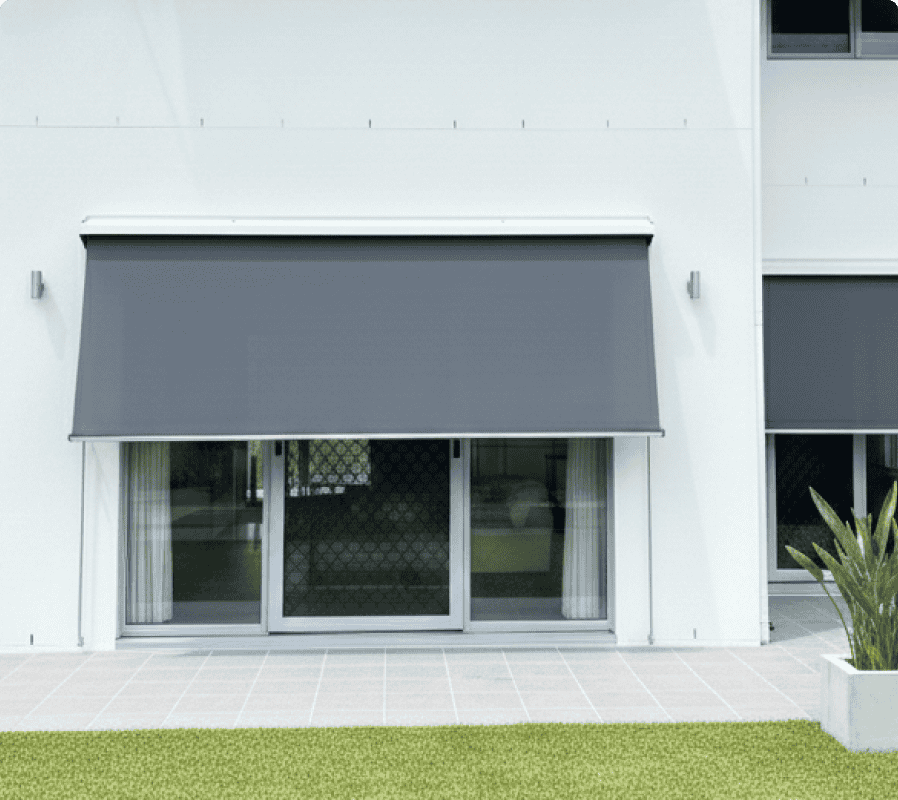 A modern glass sliding door with a large, dark grey exterior roller blind partially lowered, set in a white wall, with tiled patio and green grass in front.
