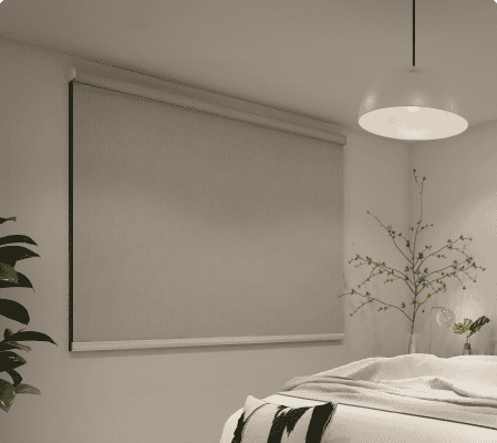 A bedroom with a closed roller blind on the window, a bed with white bedding, a hanging light fixture, and plants in the corners.