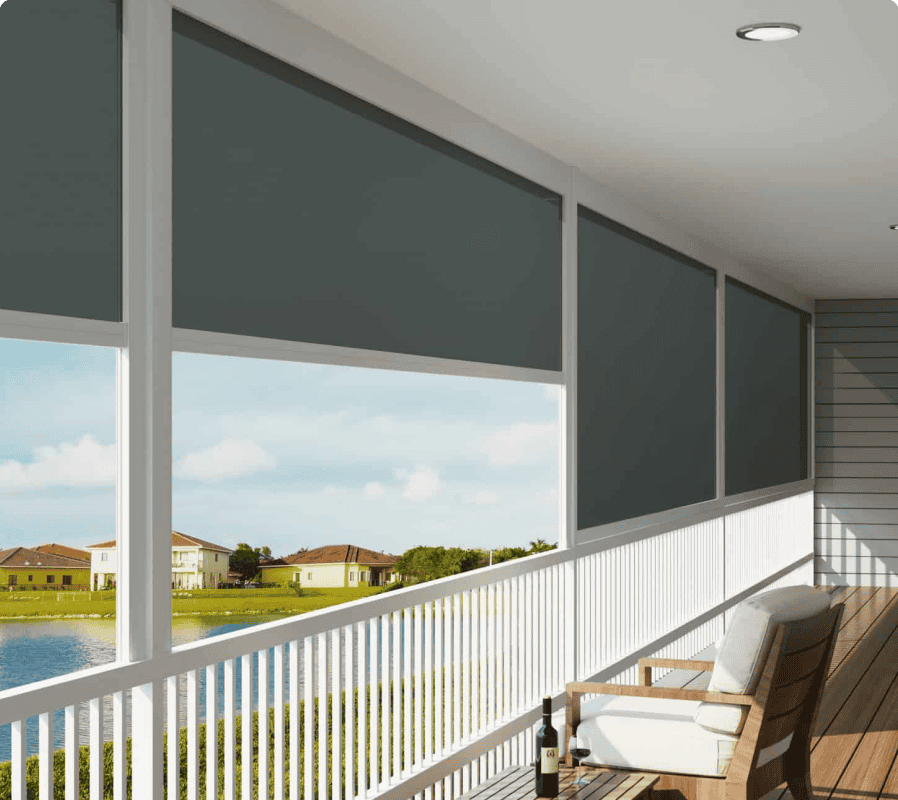 A covered porch with outdoor roller shades, a white railing, a cushioned bench, and a small table with wine, overlooking a pond and houses in the distance.