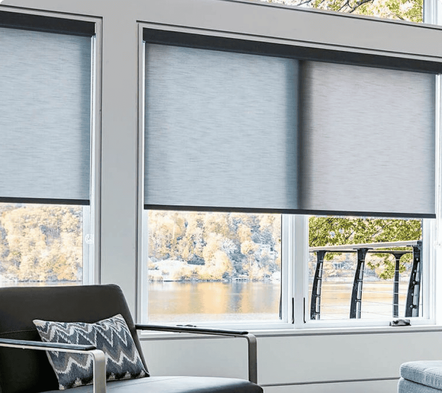 Three large windows with gray roller shades partially down overlook a lake with trees. A modern chair with a patterned pillow sits in the foreground.