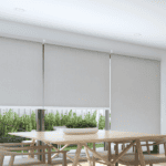 Modern dining area with a wooden table and chairs next to large windows covered by light-colored roller blinds; greenery is visible outside.