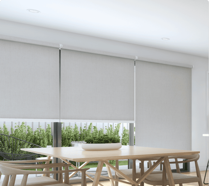 Modern dining area with a wooden table and chairs next to large windows covered by light-colored roller blinds; greenery is visible outside.