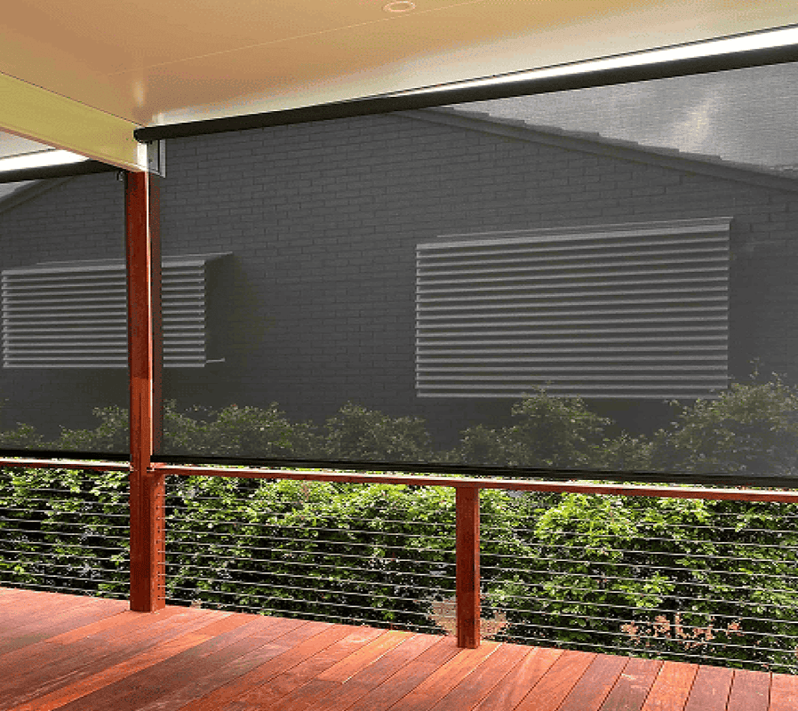 A covered patio with wooden flooring and cable railing features a semi-transparent outdoor roller blind, partially revealing a nearby brick building and greenery outside.