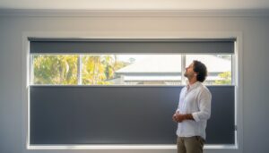 A man in a white shirt stands indoors looking at large windows with partially drawn gray blinds; trees and a house are visible outside.