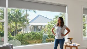 A woman stands by a large window with a gray roller blind, smiling, with a drill and screws on a small table beside her; a house and trees are visible outside.