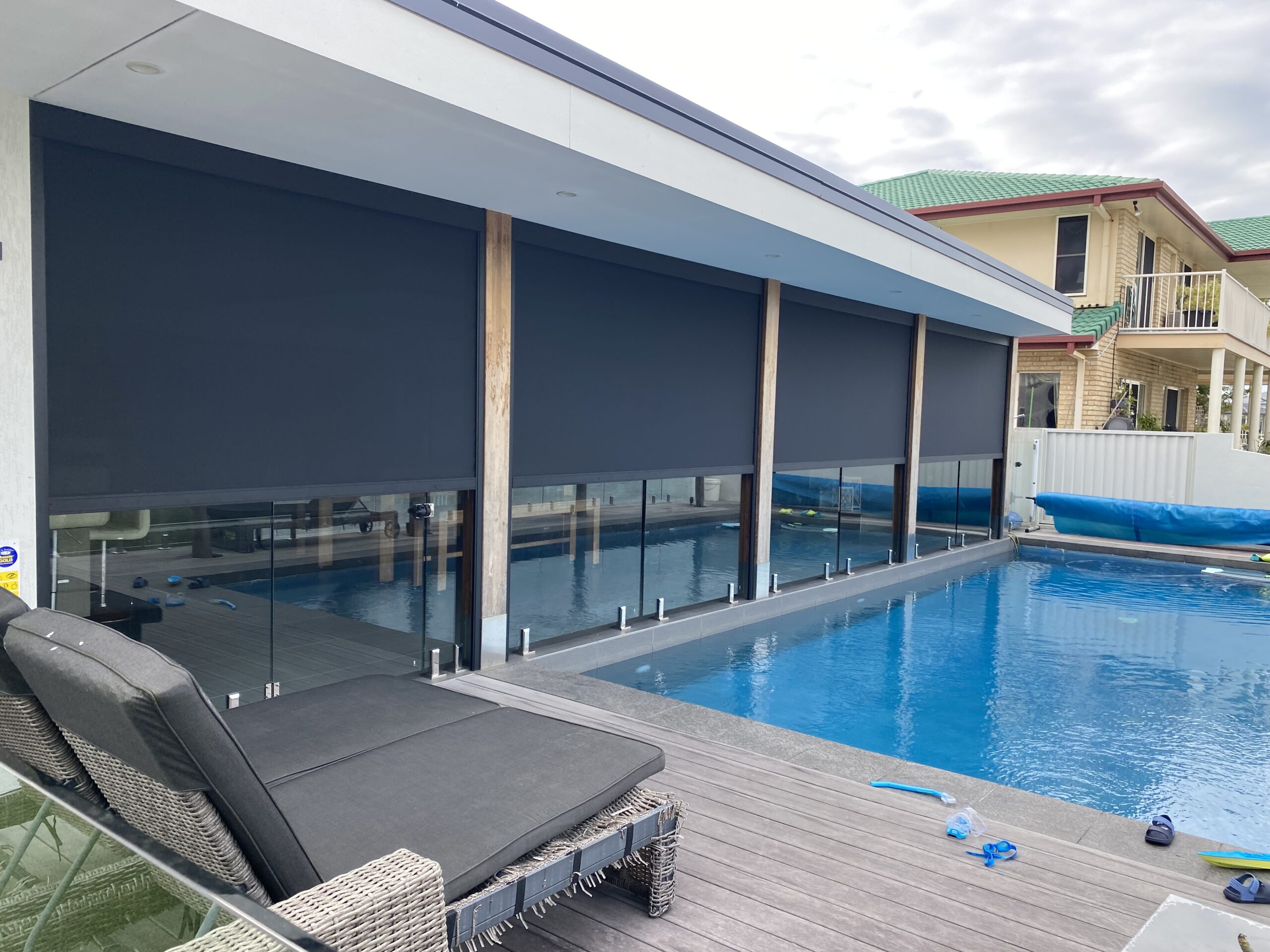 A swimming pool with an adjacent terrace featuring a grey lounger and a house exterior with extended black outdoor blinds.