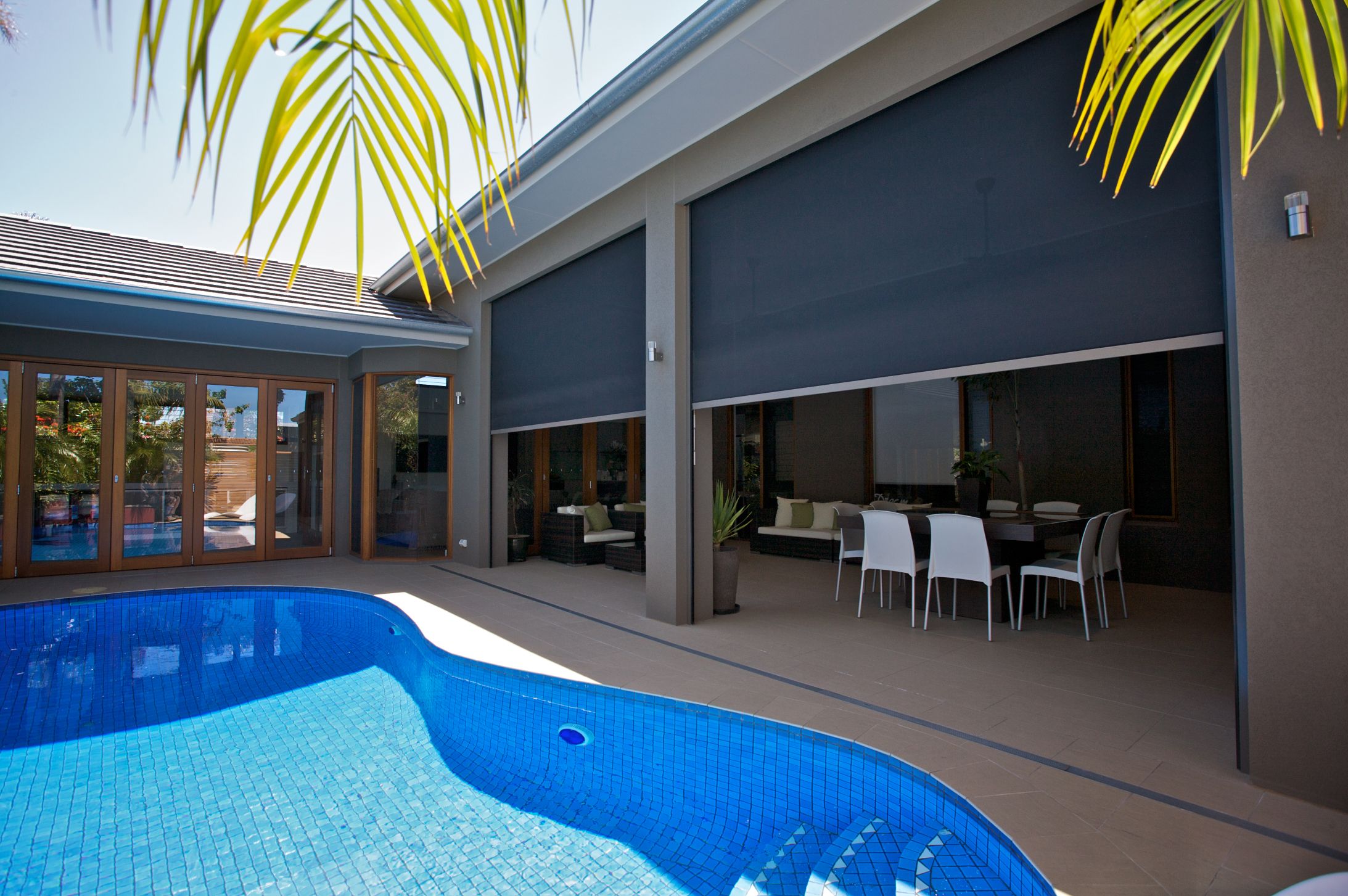 A blue swimming pool next to a covered patio featuring a dining set and lounge furniture enclosed by retractable privacy screens.